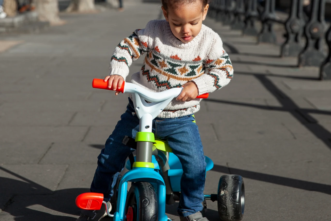 القلاف_للدراجات_النارية_ طفل صغير يرتدي سترة منقوشة وبنطلون جينز يركب دراجة ثلاثية العجلات زرقاء اللون على طريق مرصوف في الهواء الطلق. _دراجة_نارية