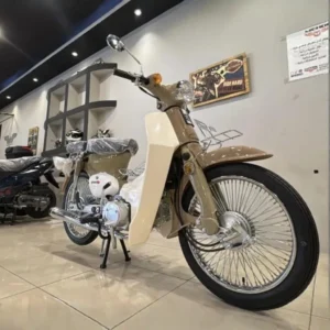 القلاف_للدراجات_النارية_ أ دراجة مهدي كلاسيكية كلاسيكية خرينق 110 جـ شبية شبية هوندا 70 بلون كلاسيكي من القلاف لدراجات نارية معروضة داخل صالة بأرضية لامعة وتظهر بجانبها دراجات نارية وملصقات في الخلفية. _دراجة_نارية